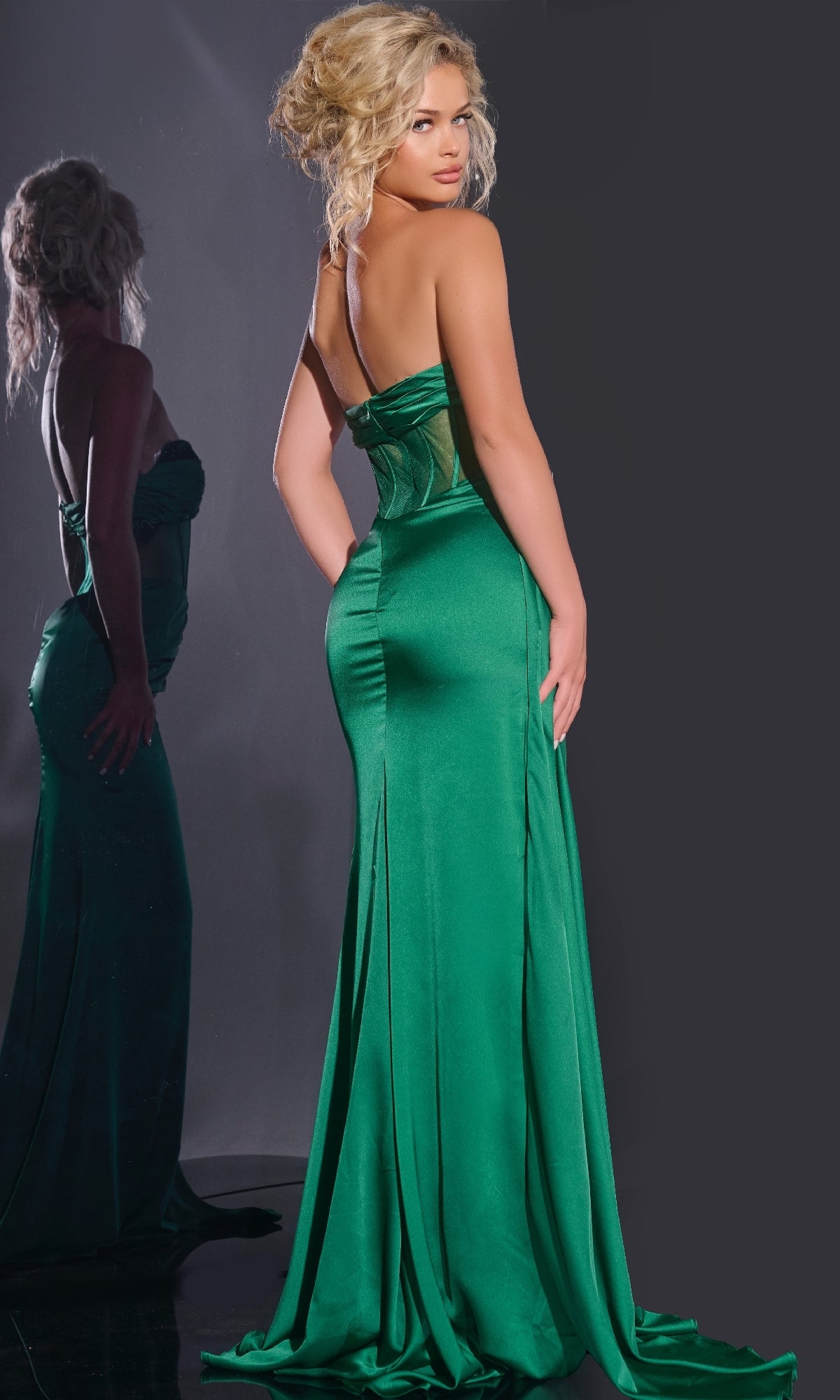 A blonde woman with her hair in an updo wears the Jovani 43157 long green satin prom dress featuring a sheer corset bodice and backless design, standing before a mirror with her reflection visible.